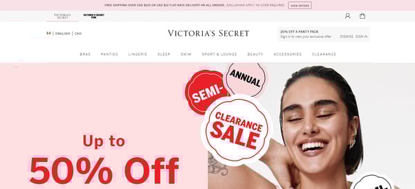 Die Hauptseite der Victoria's Secret-Website mit dem Menü mit verschiedenen Produktkategorien von Kosmetika bis Accessoires und dem Banner, das Ausverkaufsaktionen mit Rabatten auf verschiedene Produkte ankündigt. Die Webseite des Einzelh?ndlers ist in den Farbt?nen Rosa, Wei? und Schwarz gehalten und tr?gt das Victoria's Secret-Logo oben auf der Seite.